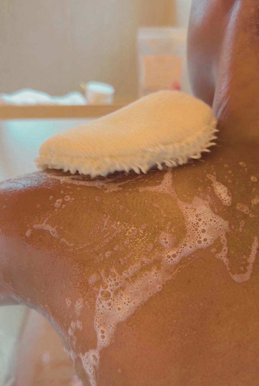 Person using Löwin Crystal cleansing bath gel and a scrubber on their back with soap suds in a bathroom setting